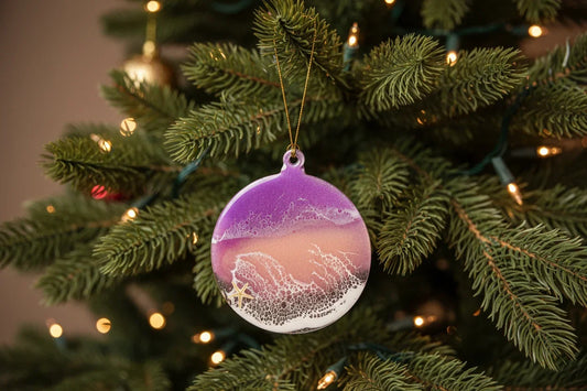 A round beach-themed Christmas ornament hanging on a decorated Christmas tree. The ornament features a purple-to-pink gradient sky, white waves, and a small starfish at the bottom, with a gold hanging string.
