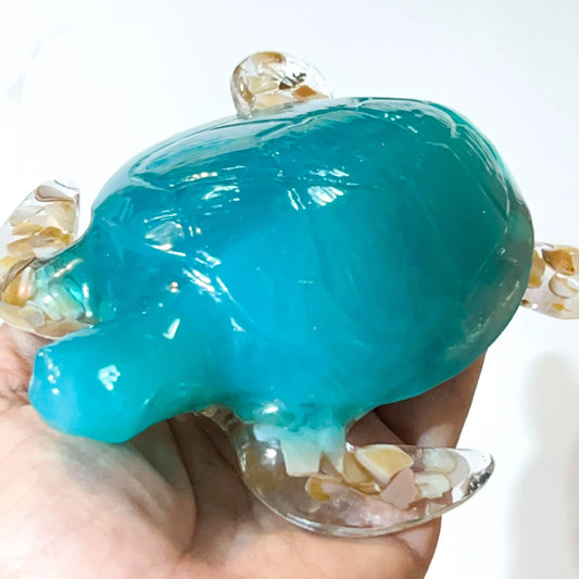 A small decorative turtle figurine with a glossy turquoise-blue shell and clear marbled legs and head, being held in a person's hand against a plain white background.