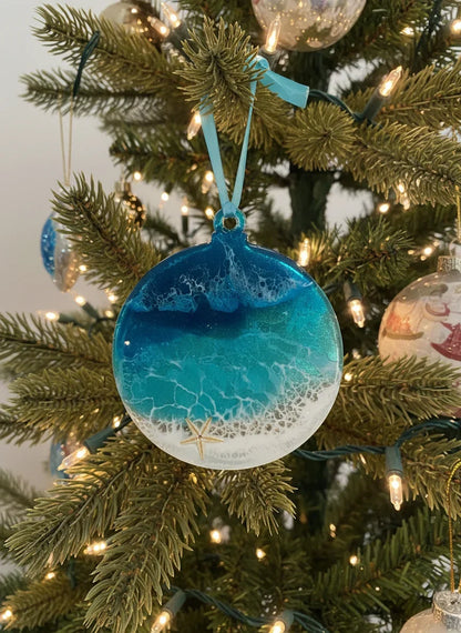 A round beach-themed Christmas ornament with a blue and white ocean wave design hangs on a decorated Christmas tree with warm lights.