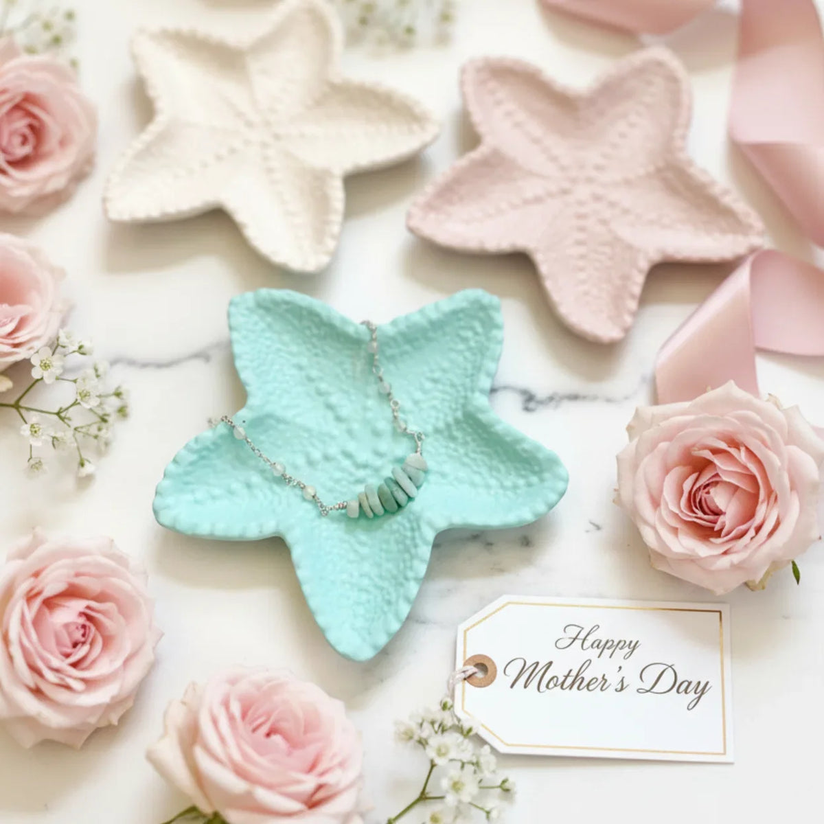Four textured starfish-shaped jewelry display stands in pastel colors (cream, light pink, mint green, and pale pink) arranged on a white marble surface with pink roses, baby's breath, and a 'Happy Mother's Day' tag.
