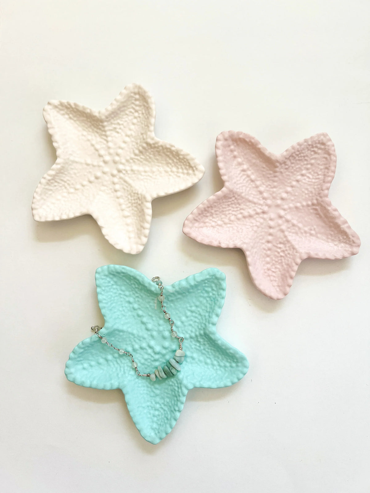 Three textured starfish-shaped jewelry trays in cream, light pink, and mint green, arranged on a white background with a silver beaded necklace resting on the mint green tray.