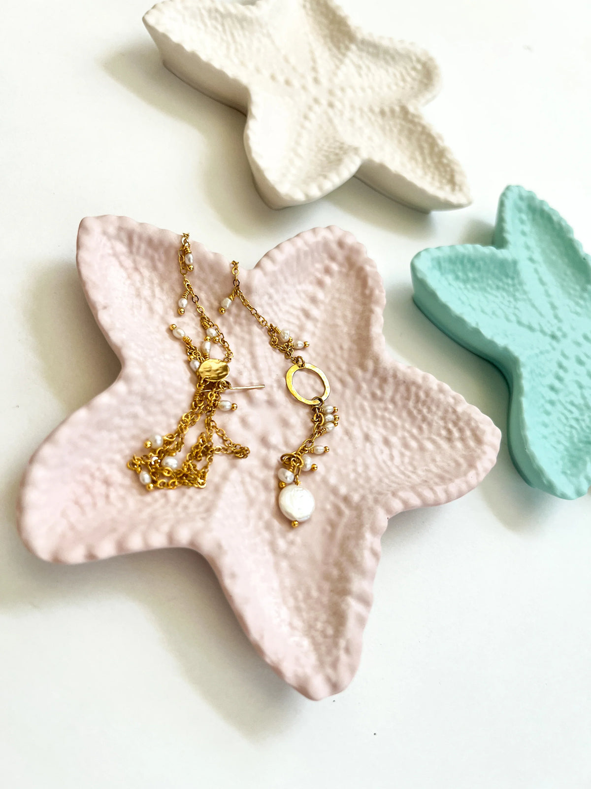 Gold chain necklace and matching earrings with white pearl-like beads displayed on a pink starfish-shaped tray, with additional decorative starfish trays in white and mint green on a white background.