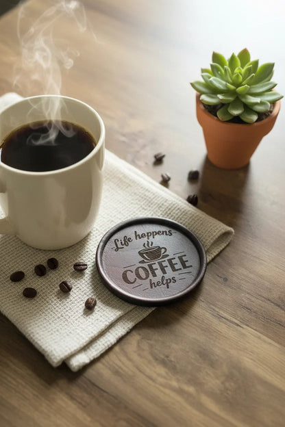 A round dark coaster with a coffee cup graphic and the words 'Life happens COFFEE helps' sits on a beige cloth beside a cup of black coffee on a wooden table, with coffee beans scattered around and a small potted succulent in the background.