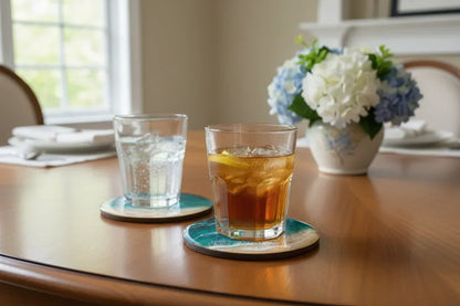 Two clear drinking glasses resting on round teal coasters on a wooden dining table, with one glass containing an amber drink and a lemon slice. A vase of flowers and a softly blurred indoor room are in the background.