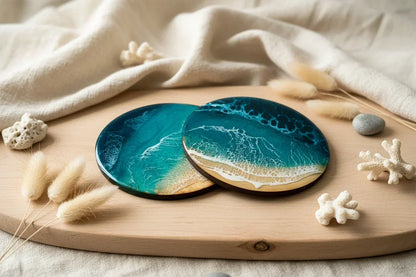 Two round coasters with an ocean wave design in teal, blue, and beige, displayed on a wooden board with decorative dried grass and coral pieces.