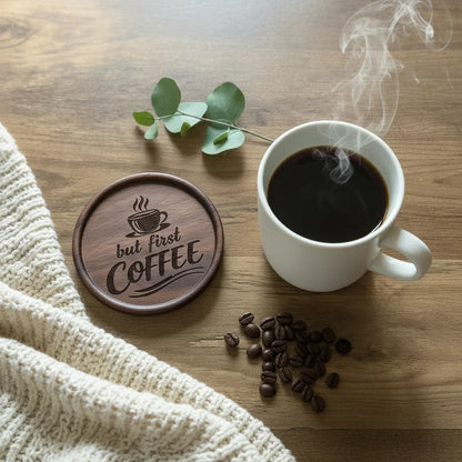 White mug with steaming coffee, wooden coaster with 'but first, COFFEE' text, coffee beans, and eucalyptus leaves on a wooden surface.