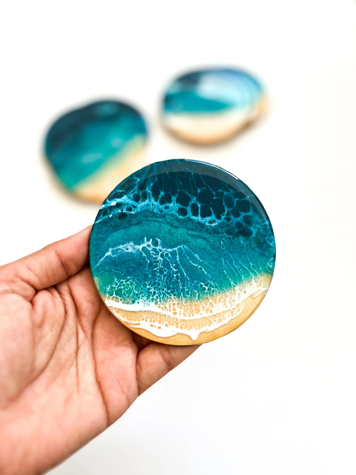 A hand holds a round resin coaster featuring an ocean wave pattern in teal, blue, and white with a sandy beige shore. Two more coasters are blurred in the background on a white surface.
